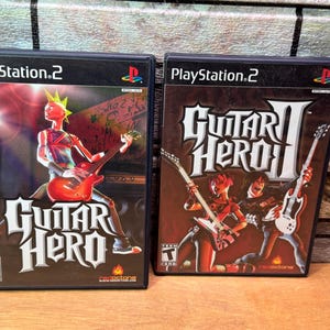 May include: Two PlayStation 2 video game cases. The first is "Guitar Hero" with a figure playing a red guitar. The second is "Guitar Hero II" with figures playing guitars. Both cases are dark with white text.
