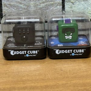 Op de afbeelding: Twee Fidget Cubes in een doorzichtige plastic verpakking. Eén is zwart met knoppen en een joystick, de andere is groen met een Hulk-gezicht. Beide zijn voorzien van de tekst "Fidget Cube" en "antsy labs".