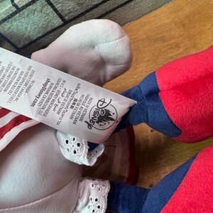 May include: A plush doll with red, white, and blue accents. The doll has a white head, red arms and legs, and blue sleeves. The doll's clothing includes a white dress with red stripes and white lace trim. The Disney logo is visible.