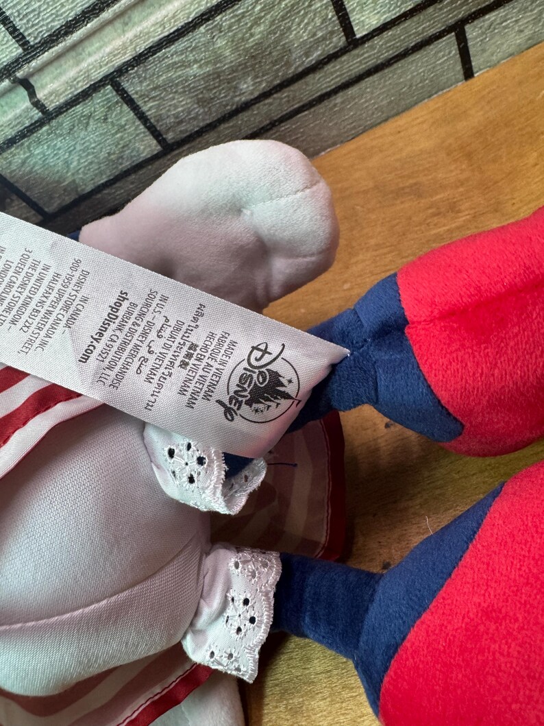 May include: A plush doll with red, white, and blue accents. The doll has a white head, red arms and legs, and blue sleeves. The doll's clothing includes a white dress with red stripes and white lace trim. The Disney logo is visible.