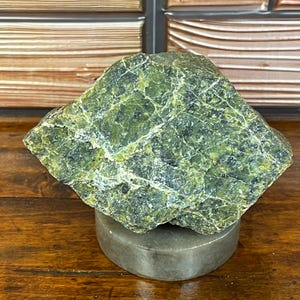 May include: A rough, angular specimen of green and black serpentine mineral resting on a smooth, circular, light grey stone base. The rock features a complex, mottled texture with visible white veining running across its uneven, crystalline surface.