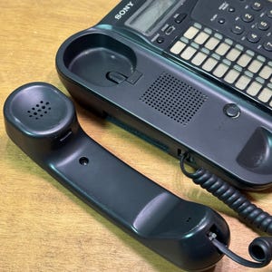 Puede incluir: Un tel&eacute;fono con cable Sony negro con el auricular separado. El tel&eacute;fono tiene un teclado, una pantalla y un altavoz. El auricular est&aacute; conectado a la base con un cable en espiral. El tel&eacute;fono est&aacute; sobre una superficie de madera.