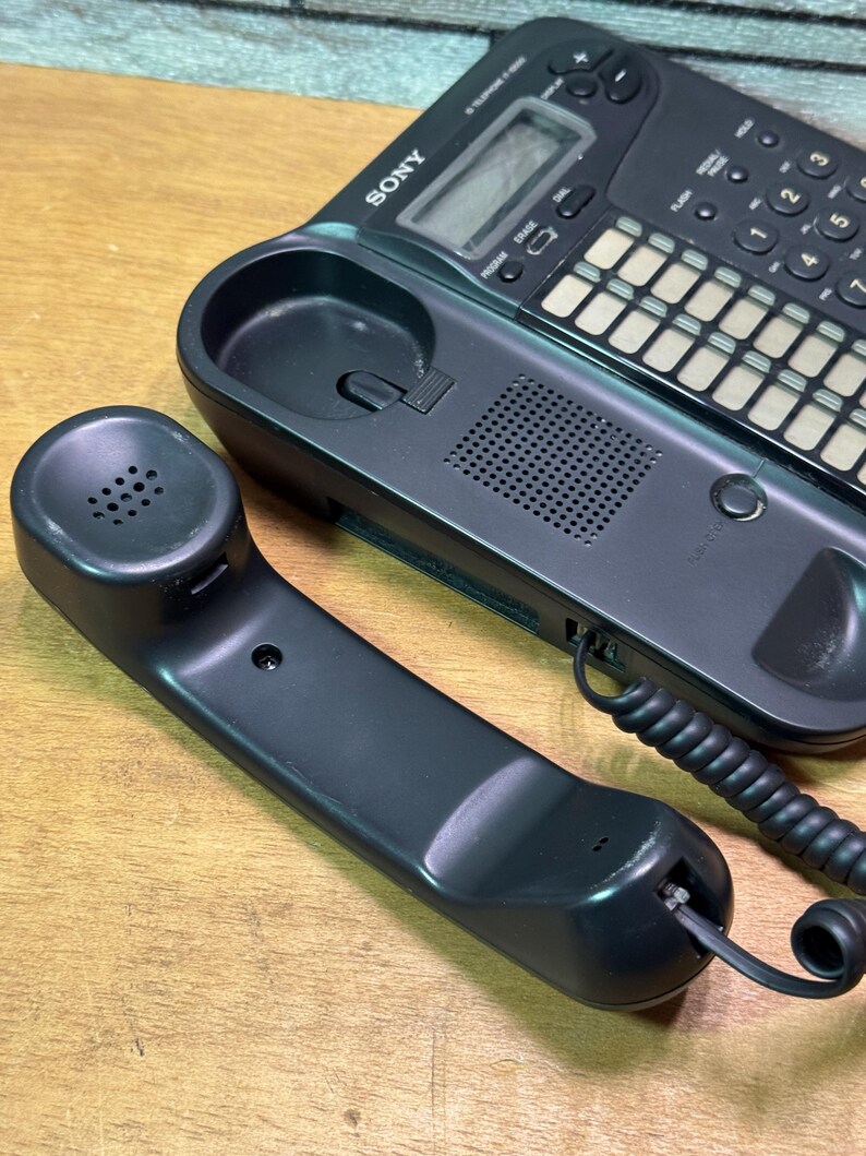 Puede incluir: Un tel&eacute;fono con cable Sony negro con el auricular separado. El tel&eacute;fono tiene un teclado, una pantalla y un altavoz. El auricular est&aacute; conectado a la base con un cable en espiral. El tel&eacute;fono est&aacute; sobre una superficie de madera.