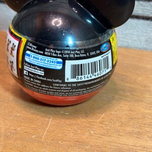 May include: A black and red Mickey Mouse-shaped container with attached ears. The container has a barcode and text that includes the Disney and Just Play logos. The container is on a wooden surface.