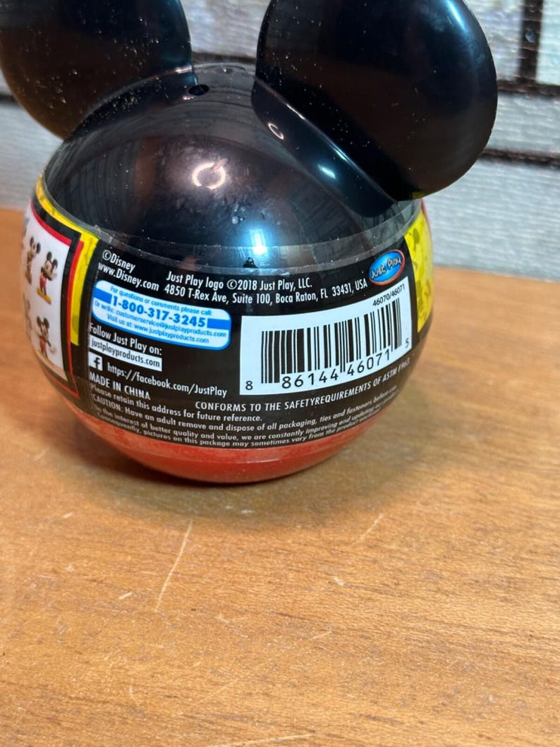 May include: A black and red Mickey Mouse-shaped container with attached ears. The container has a barcode and text that includes the Disney and Just Play logos. The container is on a wooden surface.