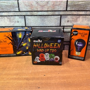 May include: Three Halloween-themed boxes on a wooden surface. The boxes contain a light set, wind-up toys, and a black light bulb. The light set box is orange and black with a tree silhouette. The wind-up toy box is black with Halloween characters. The black light bulb box is orange and black.