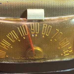 K&ouml;nnte beinhalten: Nahaufnahme eines Vintage-Radio-Zifferblatts mit einem vergilbten Kunststoffgesicht und einem goldfarbenen Zeiger. Das Zifferblatt ist mit Zahlen von 5 bis 100 gekennzeichnet. Der Text "Emerson Radio & Phonograph Corporation, N.Y. U.S.A." ist unten sichtbar.