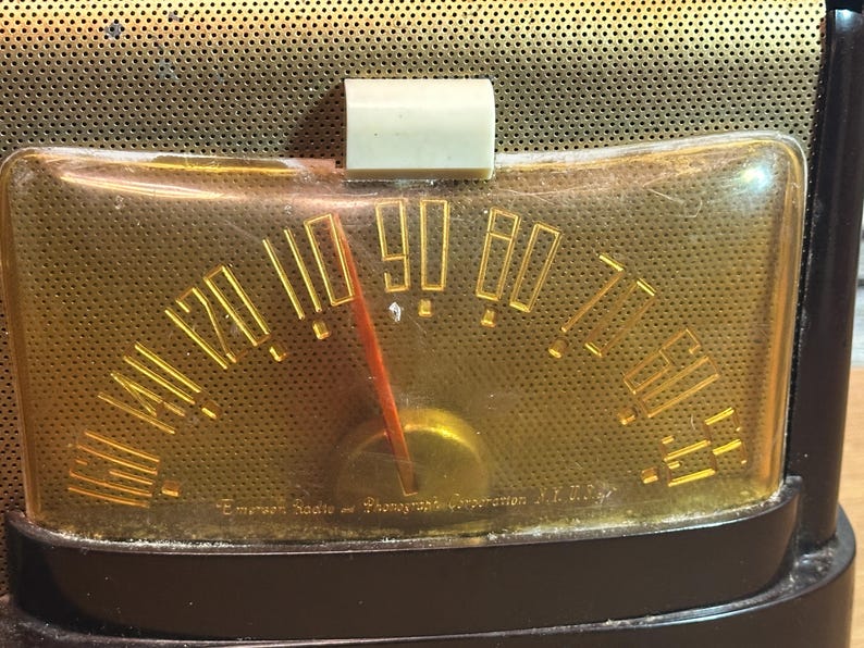 K&ouml;nnte beinhalten: Nahaufnahme eines Vintage-Radio-Zifferblatts mit einem vergilbten Kunststoffgesicht und einem goldfarbenen Zeiger. Das Zifferblatt ist mit Zahlen von 5 bis 100 gekennzeichnet. Der Text "Emerson Radio & Phonograph Corporation, N.Y. U.S.A." ist unten sichtbar.