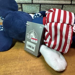 May include: A plush toy featuring a navy blue head and ears, a red and white striped hat, and a white foot. The toy has a Disney tag attached. The tag indicates the toy is suitable for ages 0+.