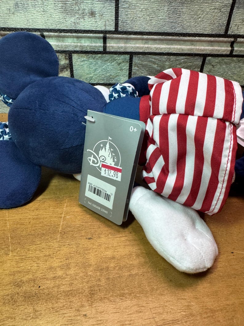 May include: A plush toy featuring a navy blue head and ears, a red and white striped hat, and a white foot. The toy has a Disney tag attached. The tag indicates the toy is suitable for ages 0+.