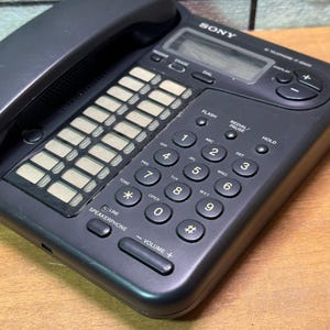 Puede incluir: Un tel&eacute;fono negro Sony IT-8000 con auricular, teclado y pantalla. El teclado tiene botones num&eacute;ricos y de funci&oacute;n, incluyendo altavoz y controles de volumen. El tel&eacute;fono est&aacute; sobre una superficie de madera.