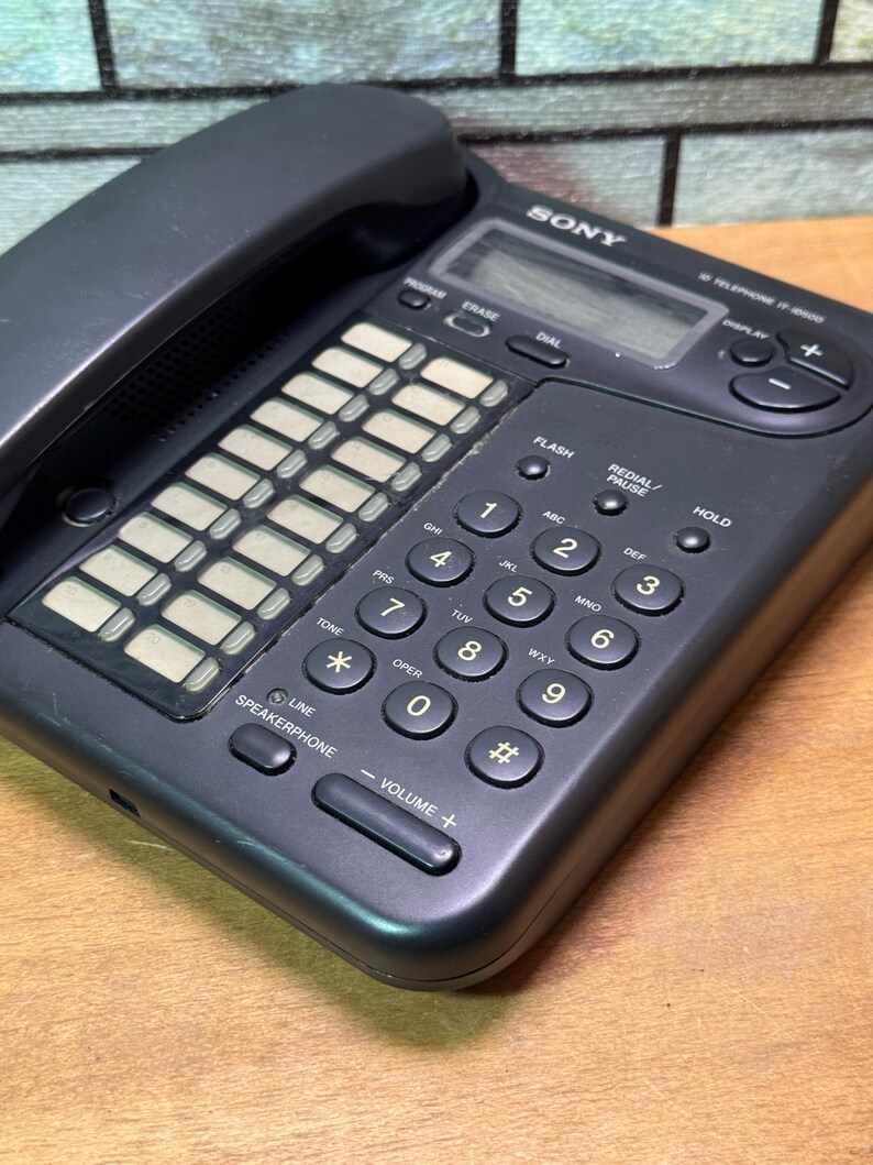 Puede incluir: Un tel&eacute;fono negro Sony IT-8000 con auricular, teclado y pantalla. El teclado tiene botones num&eacute;ricos y de funci&oacute;n, incluyendo altavoz y controles de volumen. El tel&eacute;fono est&aacute; sobre una superficie de madera.