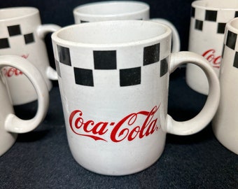 Op de afbeelding: Set van vijf witte keramische mokken met een zwart-wit geruit patroon en het Coca-Cola-logo in rood.