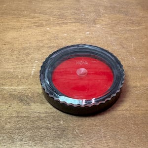 May include: A black and silver Hoya lens filter with a red interior. The filter has a circular design with a ridged edge and the brand name "HOYA" printed on the top.