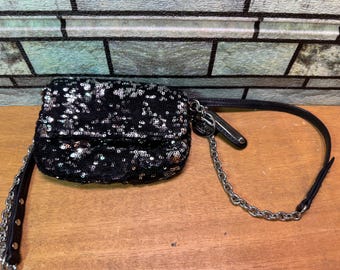 May include: Black sequined clutch with a silver chain strap and a black leather strap. The rectangular bag is covered in shiny black sequins. The chain and leather strap are attached to the bag with silver hardware.