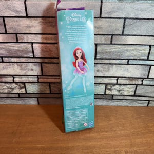 Może przedstawiać: Lalka Disney Princess Ariel w fioletowej i r&oacute;żowej sukience na niebieskim i zielonym tle. Pudełko zawiera tekst "Disney Princess" i "Imagine Ariel takes to the stage..."