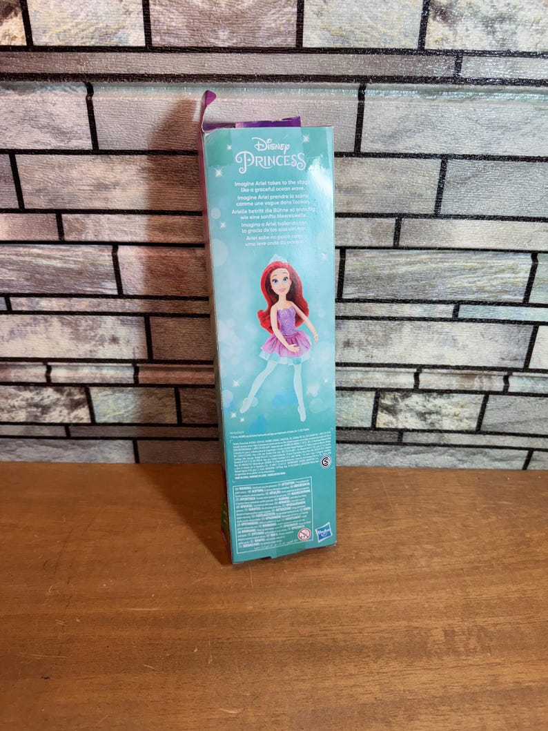 Może przedstawiać: Lalka Disney Princess Ariel w fioletowej i r&oacute;żowej sukience na niebieskim i zielonym tle. Pudełko zawiera tekst "Disney Princess" i "Imagine Ariel takes to the stage..."