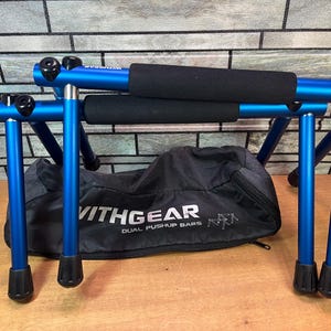 May include: A pair of bright blue metal pushup bars with black foam-padded grips and black rubber feet. The bars are positioned in front of a black zippered storage bag featuring the white text VITHGEAR DUAL PUSHUP BARS and a line drawing graphic.