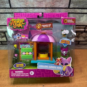 Op de afbeelding: Animal Jam Cotton Candy Hut speelset in een doorzichtige plastic verpakking. De speelset bevat een paarse en roze suikerspinstandaard met een gele basis en dierenfiguren. De verpakking heeft de tekst "Adopteer een huisdier".