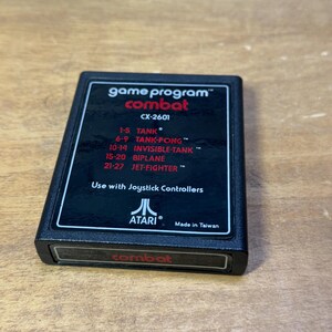 Puede incluir: Cartucho de juego Atari negro con una etiqueta roja que dice "game program™ combat CX-2601". La etiqueta enumera los diferentes modos de juego: "1-5 TANK", "6-9 TANK-PONG", "10-14 INVISIBLE-TANK", "15-20 BIPLANE", "21-27 JET-FIGHTER". La etiqueta también dice "Use with Joystick Controllers" y "ATARI® Made in Taiwan". La parte inferior del cartucho tiene una etiqueta roja que dice "combat".