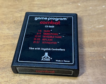May include: A black Atari game cartridge with a red label that reads "game program™ combat CX-2601". The label lists the different game modes: "1-5 TANK", "6-9 TANK-PONG", "10-14 INVISIBLE-TANK", "15-20 BIPLANE", "21-27 JET-FIGHTER". The label also says "Use with Joystick Controllers" and "ATARI® Made in Taiwan". The bottom of the cartridge has a red label that reads "combat".