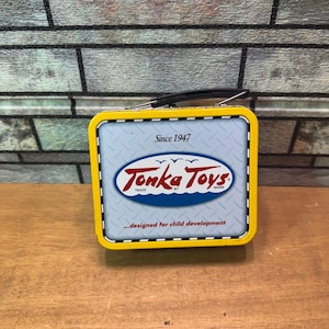 Puede incluir: Lonchera metálica vintage de Tonka Toys con marco amarillo y logotipo ovalado azul y blanco. La lonchera presenta el texto "Since 1947" y "...designed for child development."