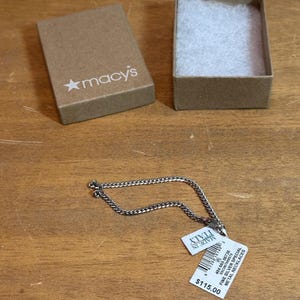May include: A silver chain necklace with a small pendant lies on a wooden surface next to an open Macy's gift box. The necklace has a tag that reads "MADE IN ITALY" and "FINE SILVER SPECIAL METAL NECKLACES".