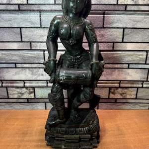 May include: A dark green statue of a female figure, adorned with intricate jewelry and a crown. The figure is holding a drum and is standing on a detailed pedestal. The statue has a weathered appearance, suggesting an antique or vintage piece.