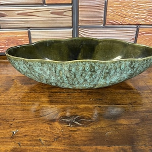 May include: An elongated oval ceramic bowl featuring a dark olive green interior and a textured, mottled exterior with shades of sage green and brown. The rim has a scalloped, wavy edge, and the overall finish appears to have a matte glaze.