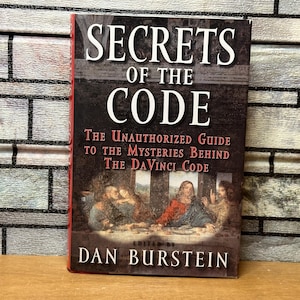 May include: A book titled "Secrets of the Code: The Unauthorized Guide to the Mysteries Behind The DaVinci Code" by Dan Burstein. The cover features a painting of people sitting at a table.