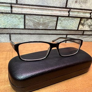 Puede incluir: Gafas de montura negra de forma rectangular, apoyadas sobre un estuche negro con textura de cuero. Las gafas tienen lentes transparentes y patillas plateadas. El fondo presenta un patr&oacute;n similar a ladrillos.