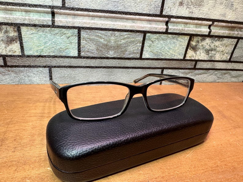 Puede incluir: Gafas de montura negra de forma rectangular, apoyadas sobre un estuche negro con textura de cuero. Las gafas tienen lentes transparentes y patillas plateadas. El fondo presenta un patr&oacute;n similar a ladrillos.