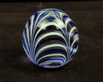 May include: A clear glass marble with a swirling pattern of white and blue stripes. The marble is spherical and sits on a dark brown surface. The stripes create a dynamic, eye-catching design.