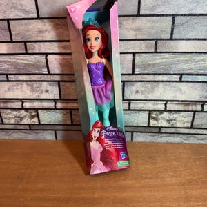Może przedstawiać: Lalka Disney Princess Ariel w fioletowej i zielonej sukience z koroną na głowie. Lalka jest zapakowana w pudełko z r&oacute;żowym i niebieskim tłem. Pudełko zawiera napis "Disney Princess" i "Obracające się ramiona księżniczki".