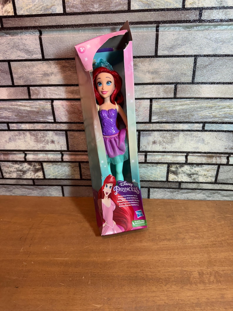 Może przedstawiać: Lalka Disney Princess Ariel w fioletowej i zielonej sukience z koroną na głowie. Lalka jest zapakowana w pudełko z r&oacute;żowym i niebieskim tłem. Pudełko zawiera napis "Disney Princess" i "Obracające się ramiona księżniczki".
