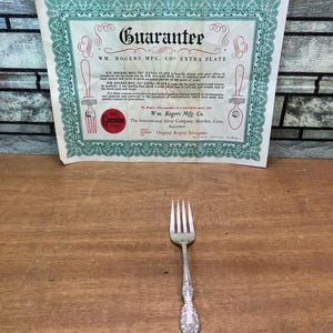 Puede incluir: Un tenedor de metal plateado con un diseño de mango floral ornamentado, acompañado de un documento de papel rectangular titulado Guarantee de Wm. Rogers Mfg. Co. Extra Plate, con bordes decorativos verdes y texto impreso en negro.