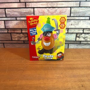 May include: A Mr. Potato Head Pirate Spud toy in its original rectangular packaging. The box features a yellow and red background with the text Mr. Potato Head, Mix 'n match fun!, and Pirate Spud. The toy includes a pirate hat, eye patch, and hook.