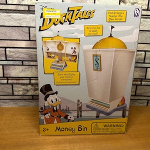 May include: A DuckTales Money Bin toy box. The box features a cartoon image of Scrooge McDuck and the text "Money Bin". The toy has a vault design with a gold dome and a dollar sign.
