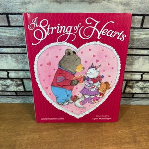 以下が含まれることがあります： ピンク色の表紙の「A String of Hearts」という本。クマがハート型のプレゼントを猫に渡しているハート型のイラストが描かれています。イラストはLynn Munsingerによるものです。