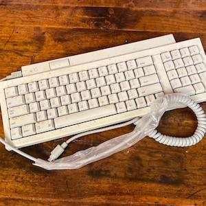 May include: A vintage off-white computer keyboard with a QWERTY layout and a numeric keypad. The keyboard features a coiled cable partially wrapped in clear plastic, a standard function key row, and a rectangular connector at the end of the cord.