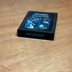 Puede incluir: Un cartucho de juego Atari negro con una imagen azul y blanca de una nave espacial y asteroides. El texto "game program ASTEROIDS" está impreso en la parte frontal del cartucho. El texto "Use with Joystick Controllers" está impreso debajo del título. El texto "CX2649" está impreso en la esquina inferior derecha. El texto "ATARI" está impreso en la esquina inferior izquierda. El texto "ASTEROIDS TM" está impreso en la parte inferior del cartucho.