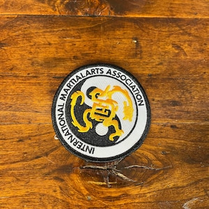 May include: A circular embroidered patch featuring a black and white yin-yang symbol at the center, overlaid with a yellow dragon design. The white border contains the black text International Martial Arts Association in a circular font.