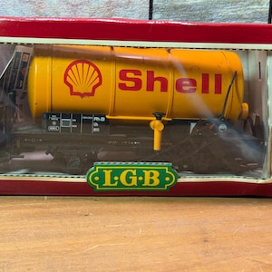 May include: A model train car in a red box. The car is yellow with the Shell logo and text. The car is on a black base with the letters "L.G.B." in green. The box has a clear plastic window.