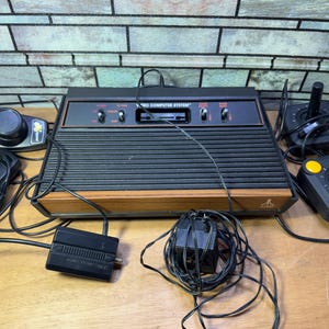 Peut inclure: Une console de jeu vidéo Atari 2600 noire avec une bordure en bois, connectée à quatre manettes. La console porte une étiquette noire et blanche qui indique "Atari Video Computer System".
