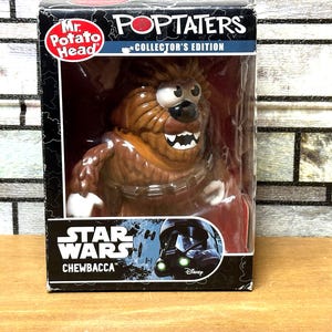 May include: A Mr. Potato Head Poptaters Collector's Edition Chewbacca figure from Star Wars inside a black rectangular box. The figure is brown and furry with white teeth, a black nose, and a bandolier. The box features the Star Wars logo.