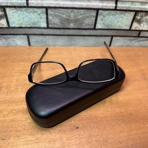 Puede incluir: Gafas con montura negra y lentes rectangulares sobre un estuche negro y texturizado para gafas. El estuche est&aacute; cerrado y tiene forma redondeada. El fondo presenta un patr&oacute;n similar a ladrillos y una superficie de madera.