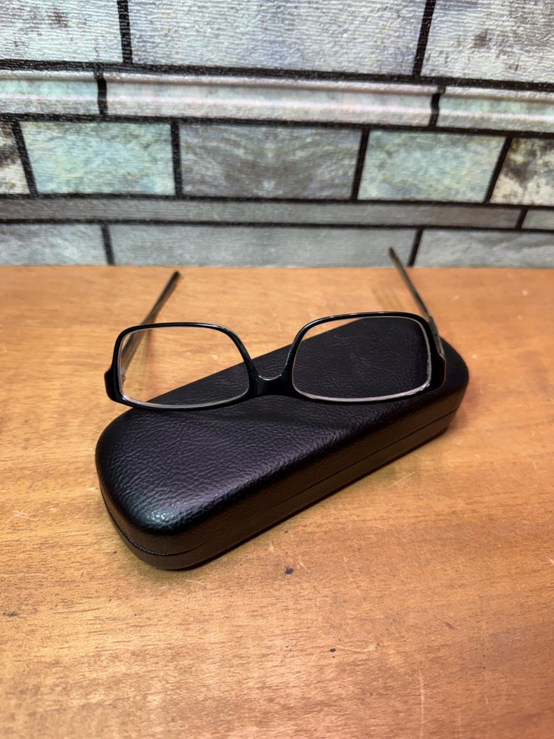 Puede incluir: Gafas con montura negra y lentes rectangulares sobre un estuche negro y texturizado para gafas. El estuche est&aacute; cerrado y tiene forma redondeada. El fondo presenta un patr&oacute;n similar a ladrillos y una superficie de madera.