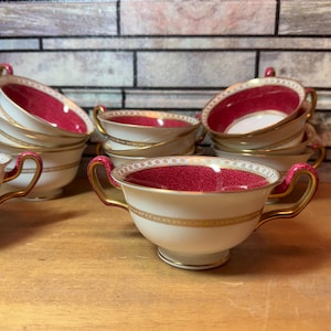 May include: A set of eight vintage teacups with a white base, pink rim, and gold trim. The cups are stacked in a pyramid shape on a wooden surface.