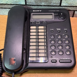 Puede incluir: Tel&eacute;fono negro Sony IT-ID600 con auricular, teclado y pantalla. El tel&eacute;fono tiene botones para marcar, flash, rellamada/pausa y retenci&oacute;n. Tambi&eacute;n son visibles los controles del altavoz y del volumen.