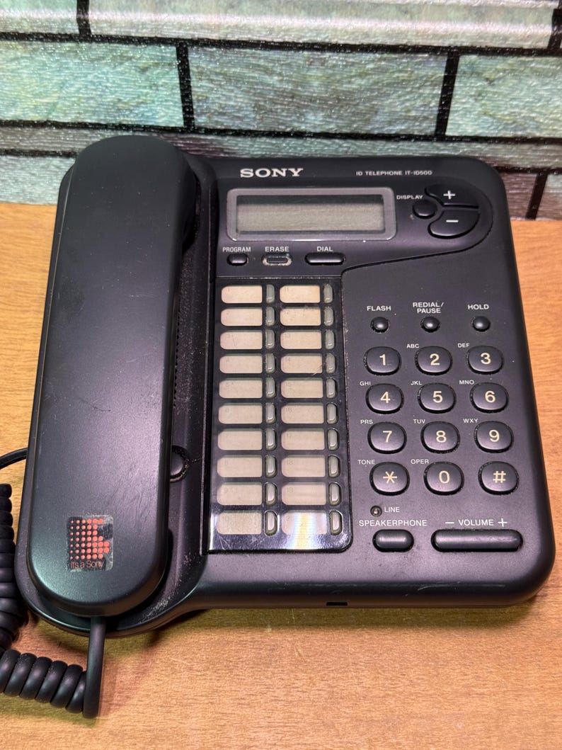 Puede incluir: Tel&eacute;fono negro Sony IT-ID600 con auricular, teclado y pantalla. El tel&eacute;fono tiene botones para marcar, flash, rellamada/pausa y retenci&oacute;n. Tambi&eacute;n son visibles los controles del altavoz y del volumen.